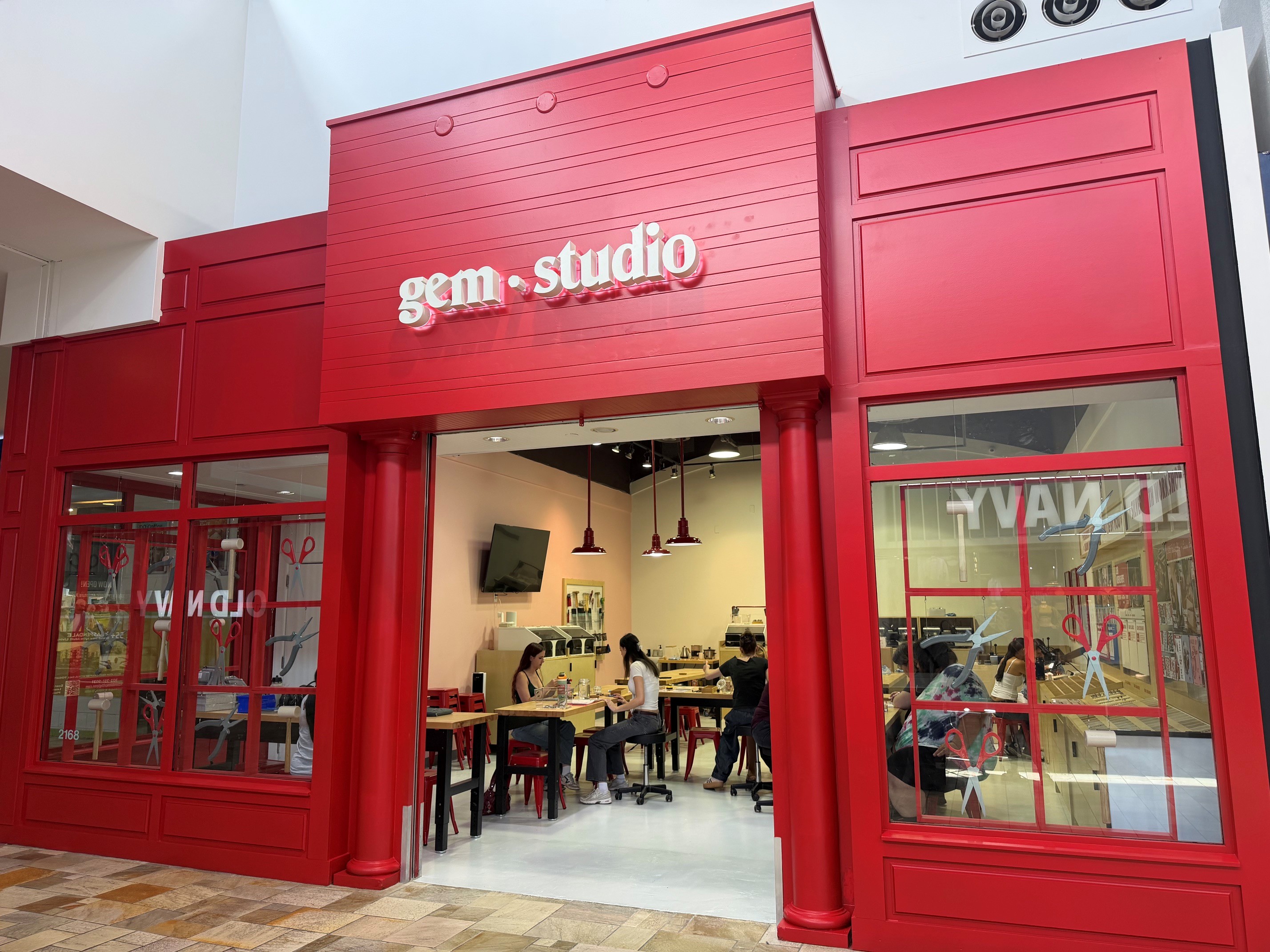 Bright red storefront for Gem Studio jewelry workshop and an open interior with people seated at tables participating in jewelry-making activities.