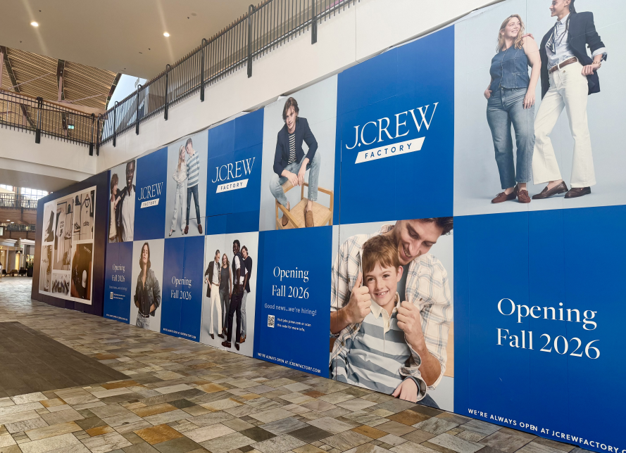 Large J.Crew Factory storefront construction wall inside a shopping mall, featuring blue promotional graphics, lifestyle photography, and text announcing “Opening Fall 2026.”