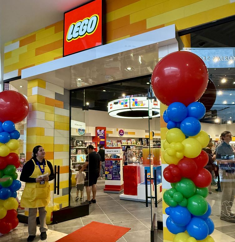 A brightly lit Lego store with balloons at the entrance and shoppers inside the store