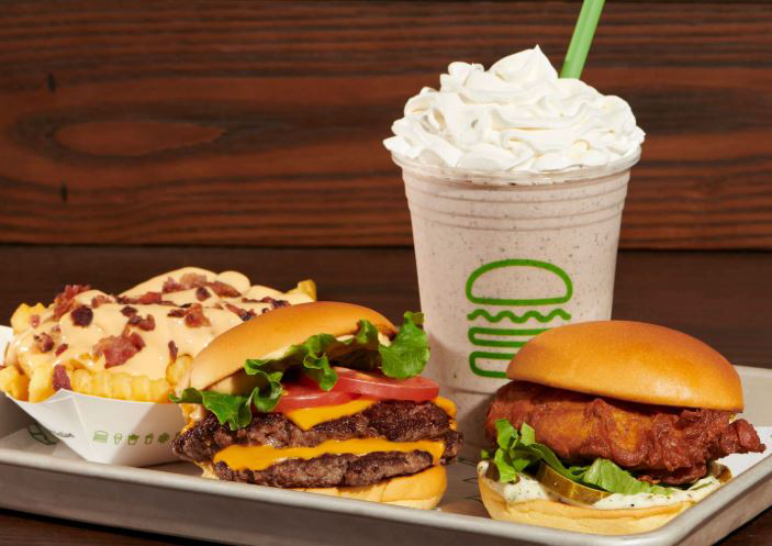 Tray with two burgers, a side of crinkle-cut fries topped with cheese sauce and bacon, and a milkshake with whipped cream 