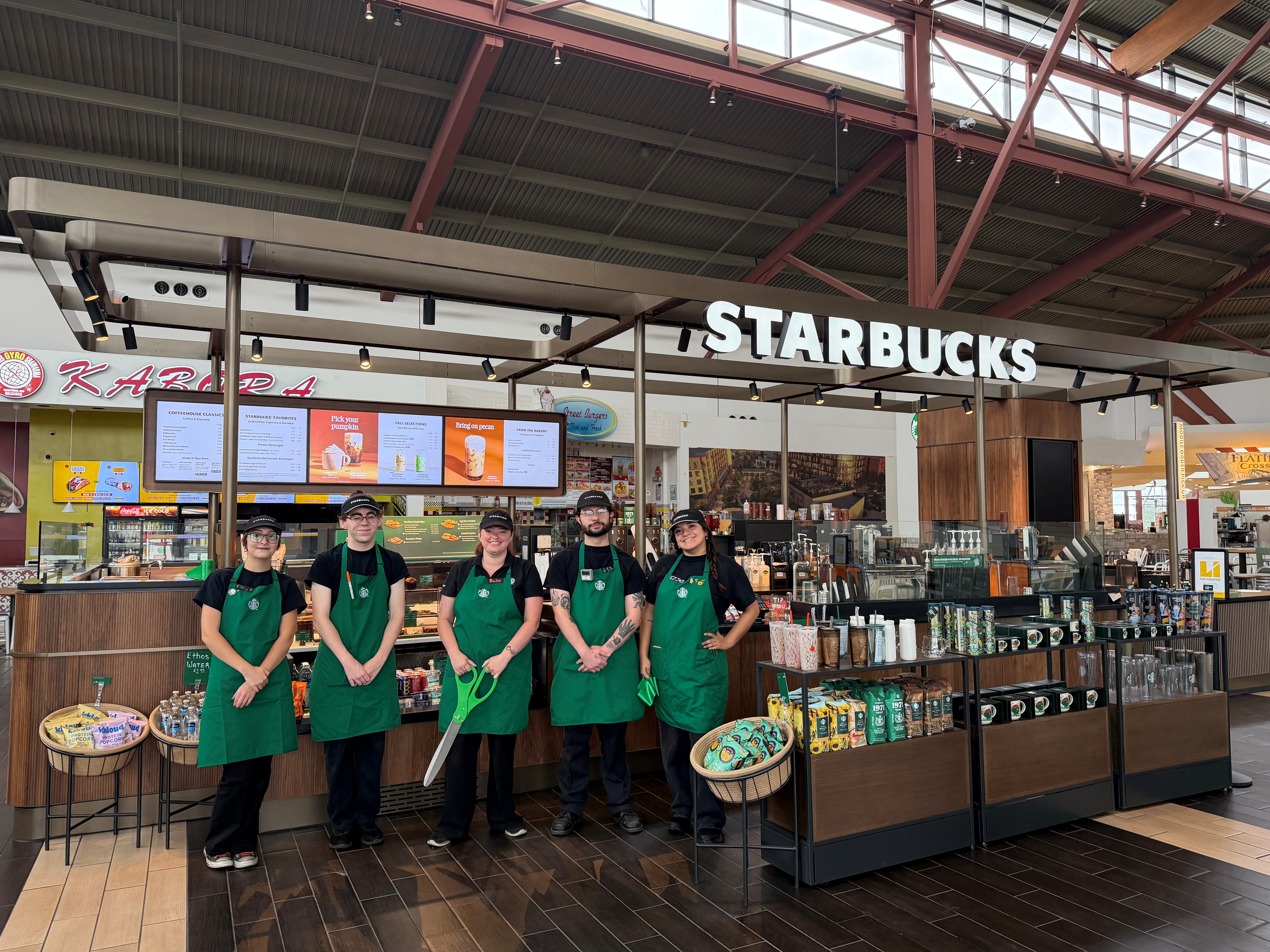 Starbucks café kiosk in a shopping mall with five team members standing in front of the counter, menu boards overhead, and packaged products displayed for sale.