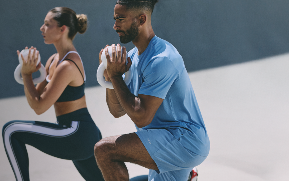 Two people perform weighted lunges during a fitness workout, each holding a kettlebell at chest height in a bright gym setting.