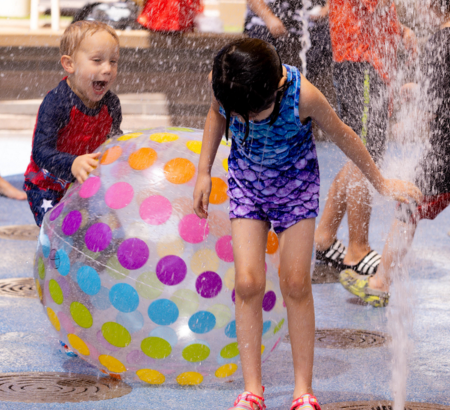 Children playing on a splash pad with a large beach ball.