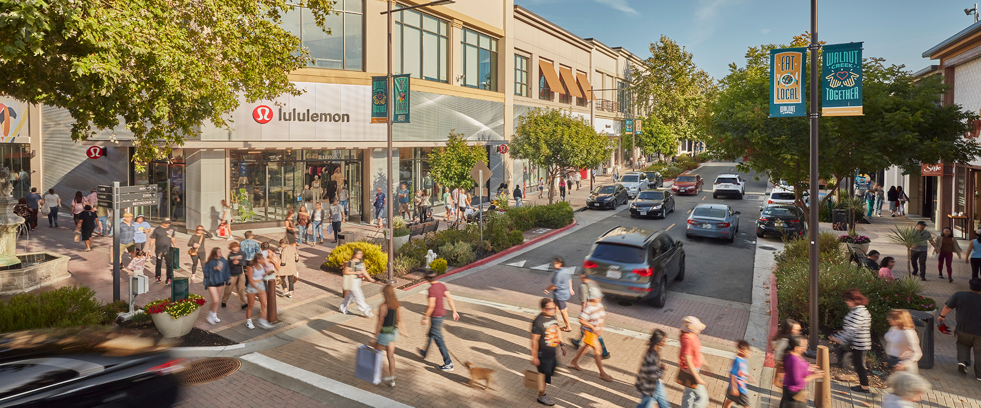 Busy outdoor shopping center in Walnut Creek with lululemon storefront, pedestrians crossing street, and cars along tree-lined retail corridor
