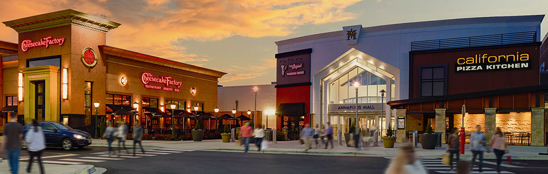 This image shows a lively outdoor shopping center at sunset. The sky is filled with warm orange and pink clouds, giving the scene a cozy, inviting feel.  In the center is the main entrance to the mall, with a tall, well-lit facade. On either side are recognizable restaurant storefronts—The Cheesecake Factory on the left and California Pizza Kitchen on the right. Both have outdoor seating areas that look active and welcoming.  There are people walking across the plaza and crosswalks, slightly blu