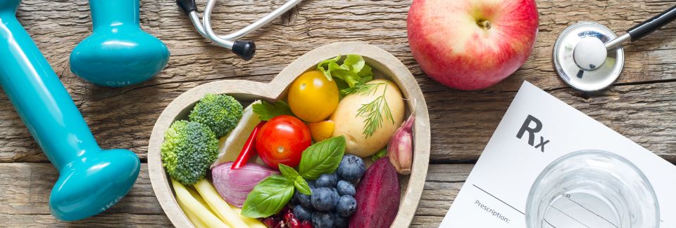Hand weights, a stethoscope, vegetables in a heart-shaped bowl, prescription page, and a glass of water.