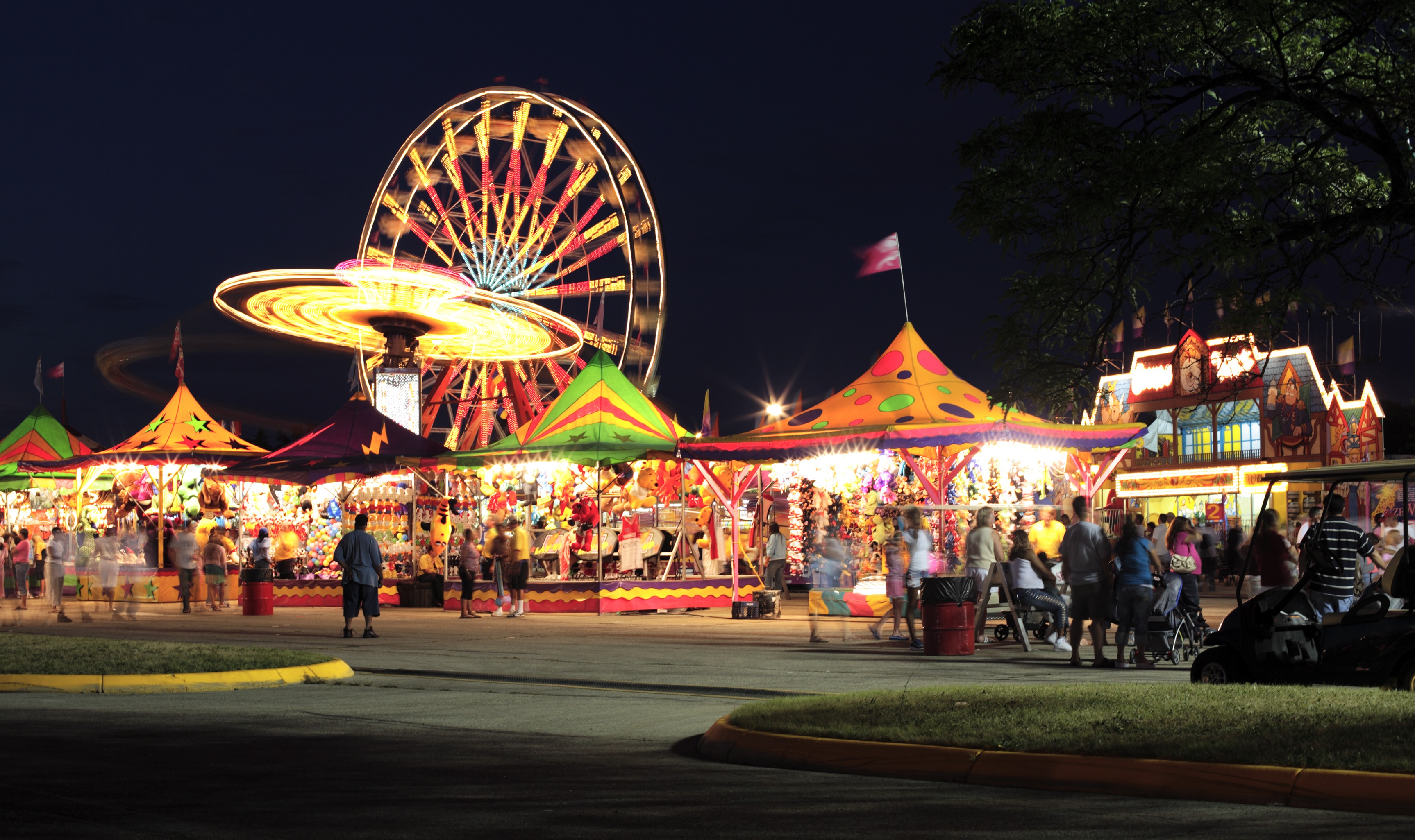 Summer carnival tents with a lit up Ferris wheel and rides. Carnival crowd blurred out walking.