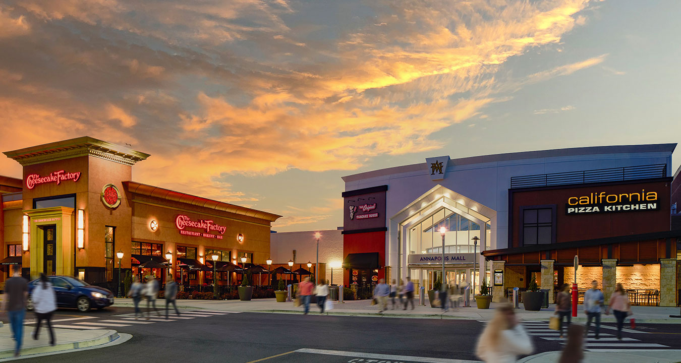 a lively outdoor shopping center at sunset. The sky is filled with warm orange and pink clouds, giving the scene a cozy, inviting feel.  In the center is the main entrance to the mall, with a tall, well-lit facade. On either side are recognizable restaurant storefronts—The Cheesecake Factory on the left and California Pizza Kitchen on the right. Both have outdoor seating areas that look active and welcoming.  There are people walking across the plaza and crosswalks, slightly blurred from motion,
