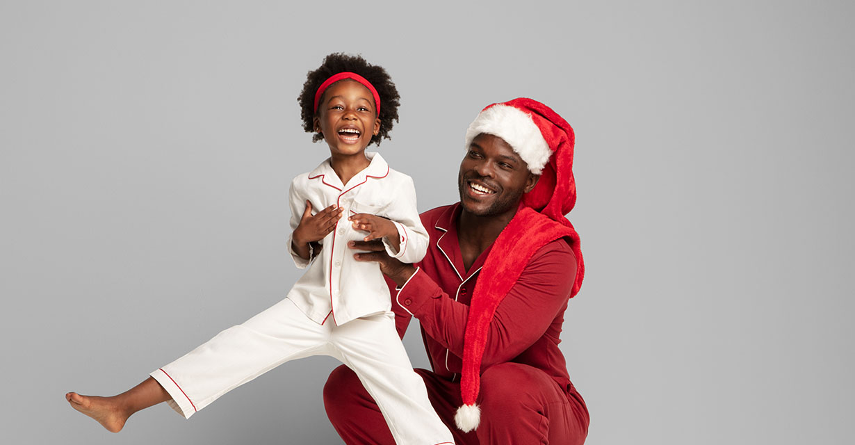 Smiling child in white pajamas with a man in red Santa pajamas against a gray background.
