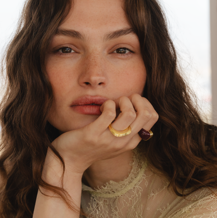 Close-up portrait of a person with wavy hair wearing gold rings and a sheer lace top, resting their hand near their face.
