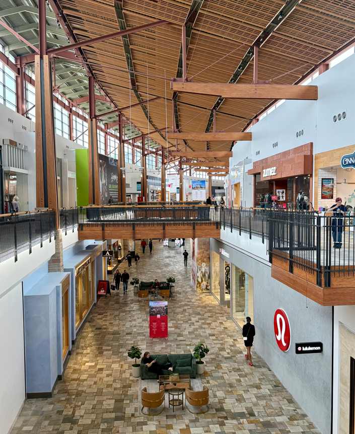 Interior of a modern two-level shopping mall with a high wood-beam ceiling, skylights, storefronts, and a few people walking and sitting below
