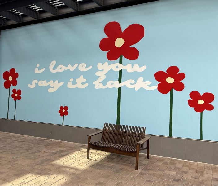 Sky-blue mural with red flowers and the message "I love you, say it back" behind a wooden bench.