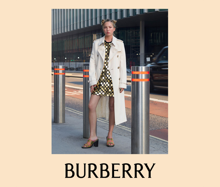 Burberry campaign image of a model in a cream trench coat over a gold mirrored-tile mini dress, standing between steel bollards on a city street.