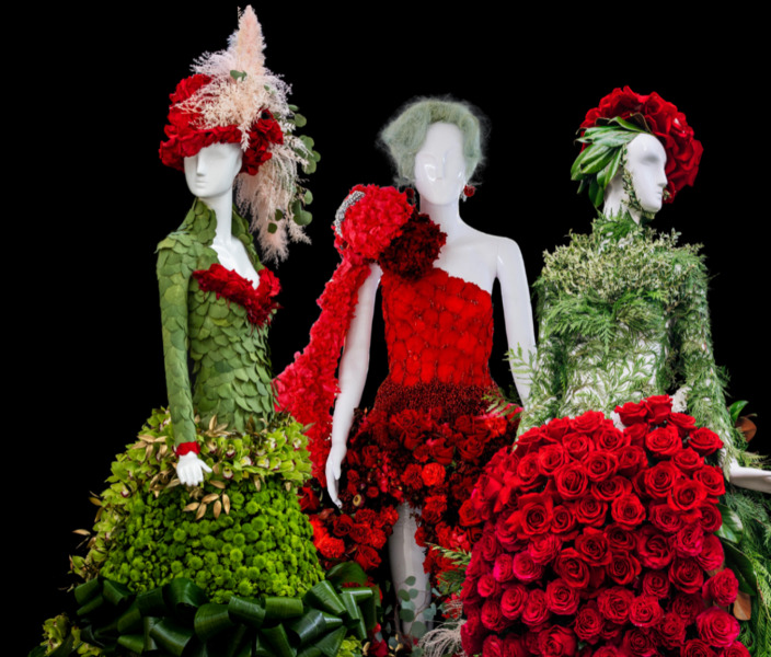 Three mannequins wear elaborate floral couture in red and green, with rose gowns, leafy designs, and dramatic floral headpieces against a black background.
