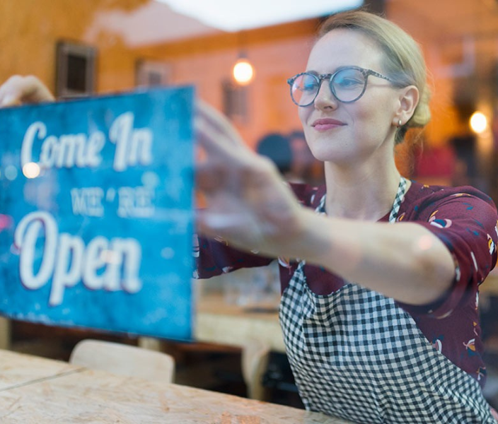 Smiling woman in glasses and a checkered apron hangs a “Come In, We’re Open” sign in a shop window, with warm lights glowing behind her.