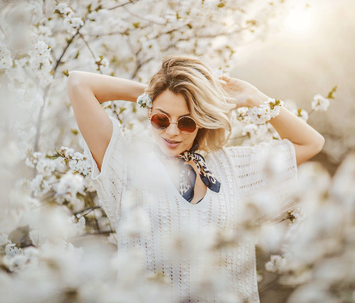 A female model wearing a neckerchief, sunglasses, and white short-sleeved sweater striking a pose in front of cherry blossoms