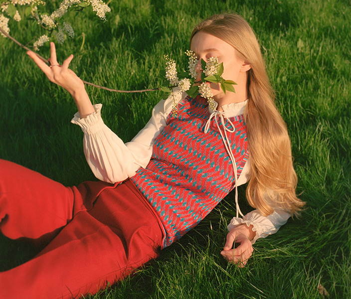 A high fashion model reclining in a field smelling a lilac and wearing a ruffled blouse, red pants, and a patterned vest
