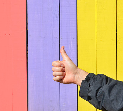 Hand giving a thumbs up in front of a multicolored fence