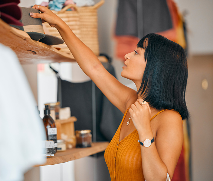 A woman in a boutique reaching for a hat displayed on a shelf