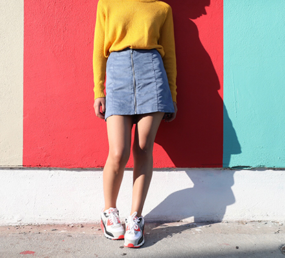 Woman wearing a skirt and sneakers standing in front of a brightly painted striped wall