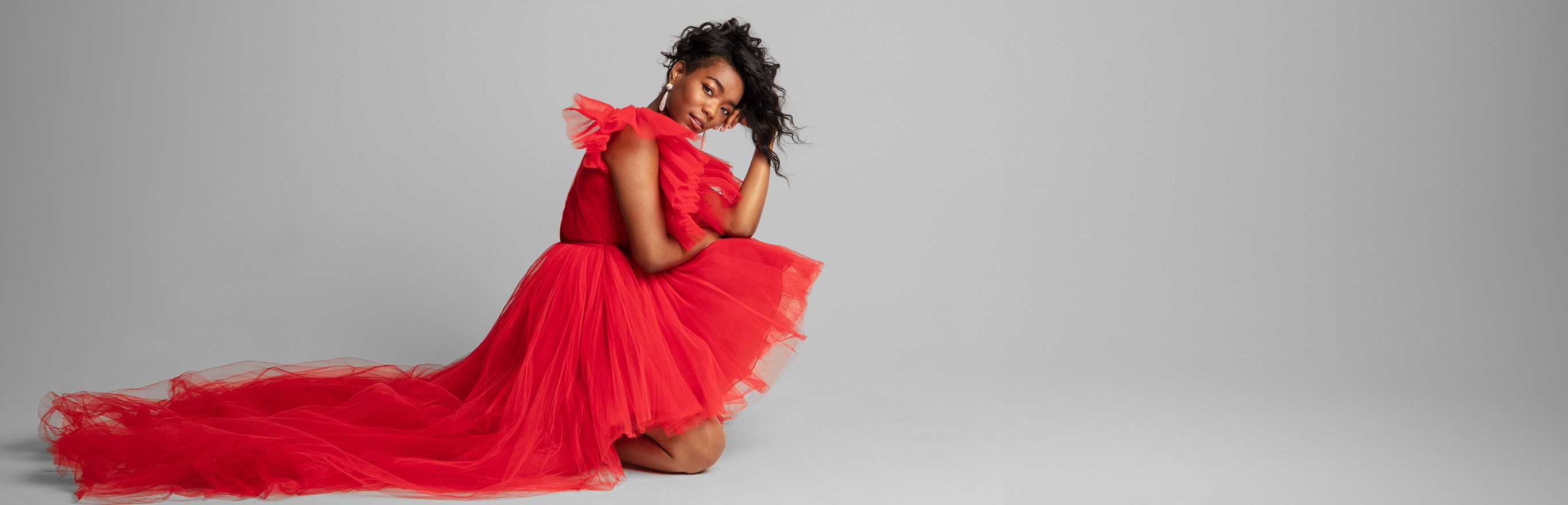 A woman in an elegant kneeling pose wearing a lacy red holiday dress with a long train.