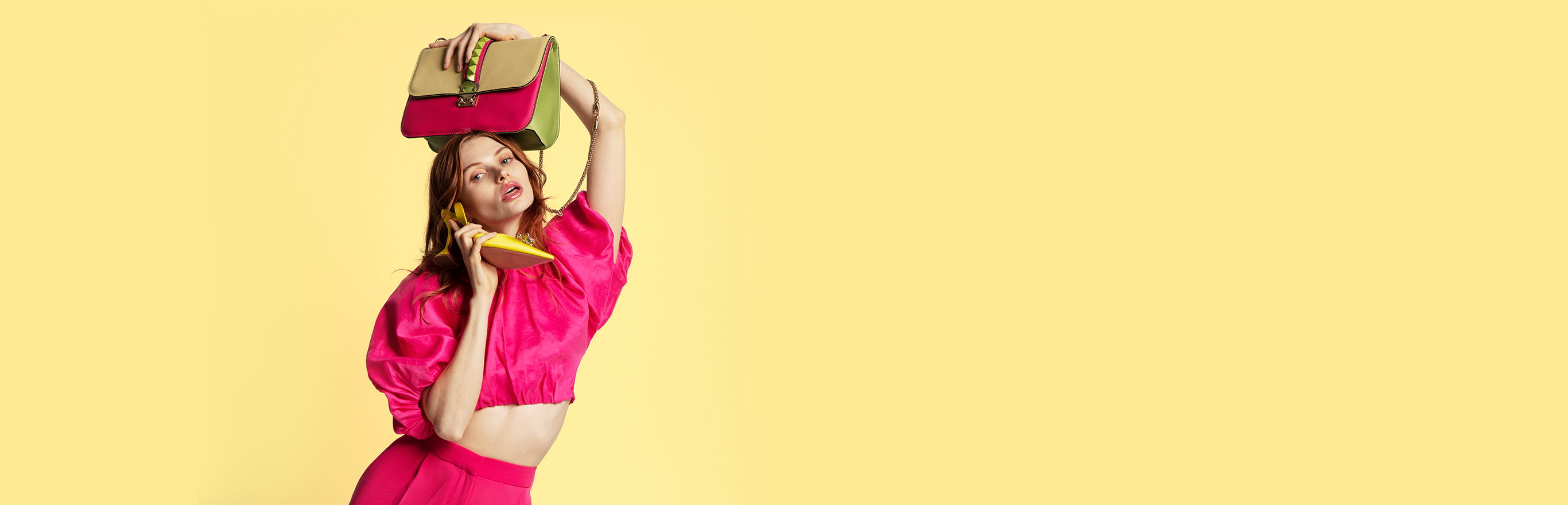 A young woman in a pink magenta crop top holding a purse over her head and speaking into a yellow high heel as a phone