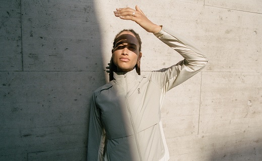 A woman in a beige ON jacket leaning against a wall with her arm up blocking the sun