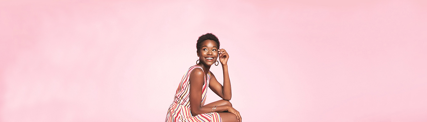 Young woman crouching down wearing a striped sundress against a pink background
