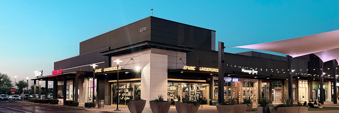 Exterior corner view of the Atomic Underground storefront in a modern outdoor shopping center at dusk, featuring black-and-white architecture, illuminated signage, large display windows, planters, and neighboring retail businesses including Massage Envy.