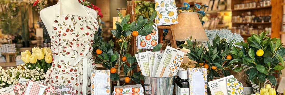 Store display featuring a mannequin wearing a strawberry-print apron, surrounded by yellow tulips, potted citrus trees with orange fruit, gingham-patterned kitchen towels and dish towels from Paper Farm Press, a lamp on a wooden ladder shelf, and shelves of home goods in the background.