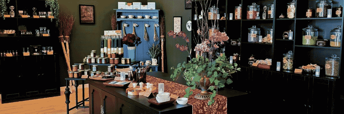 Curated boutique interior with dark shelving, glass jars, candles, dried botanicals, and a central display table featuring ceramics, flowers, and home goods.