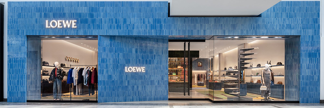 Exterior storefront of LOEWE at Fashion Square with illuminated signage, blue tiled facade, and glass display windows showcasing apparel, bags, and accessories.